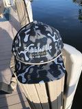 A black, gray, and white camouflage cap with 'Bubbles Up' and 'Charters GT Pro Cruiser' printed on the front, resting on a wooden outdoor chair near water.